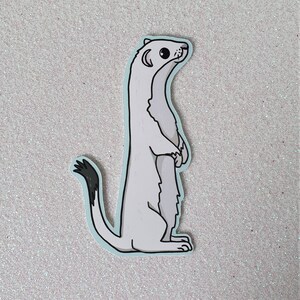 May include: A sticker featuring a cartoon ermine standing upright. The ermine is primarily white with a black-tipped tail and black accents. The sticker has a light blue border and is set against a glittery, light-colored background.