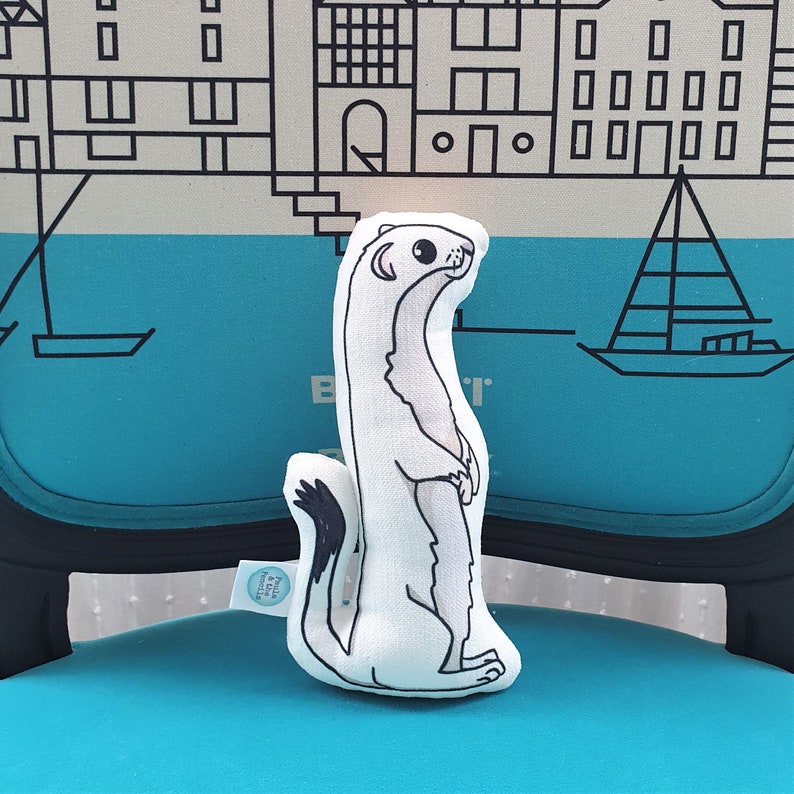 May include: White plush toy shaped like a weasel or stoat. The toy is standing upright with its paws together. The toy has black eyes and a black nose. The toy is sitting on a blue chair with a black frame.