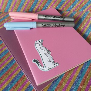 May include: A stack of pink and purple notebooks with a sticker of a white weasel. Two BIC Marking fine point permanent markers, one pink and one light blue, are on top of the notebooks.