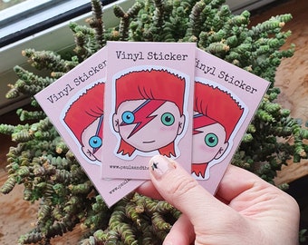May include: Three pink vinyl stickers with a cartoon illustration of David Bowie with red hair, a lightning bolt, and blue eyes. The stickers say "Vinyl Sticker" and include the website "www.paulaandthe.com".