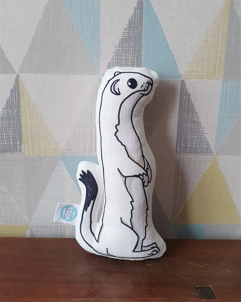 May include: White stuffed toy shaped like a weasel with black eyes and a black tail. The toy is standing upright with its paws together. The toy has a small tag on its side that says "Paula & the Pencil".