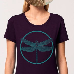 May include: A dark purple t-shirt featuring a teal dragonfly graphic within a circle. The dragonfly has intricate wings and a slender body.