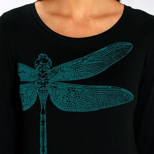 Pode incluir: Camiseta preta de manga comprida com um desenho de libélula turquesa. O rosto da pessoa é obscurecido por uma ilustração de um pássaro. A modelo está usando jeans azuis.