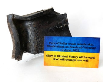 May include: A piece of a black Kalibr cruise missile skin. The missile was part of an attack on Novohrad-Volynskyi on June 9, 2022. The text on the card reads: "Glory to Ukraine! Victory will be ours! Good will triumph over evil!"