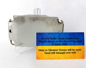 May include: A piece of a Kalibr cruise missile skin, with a card displaying text about the missile attack on Novohrad-Volynskyi on June 9, 2022. The card has the colors of the Ukrainian flag.