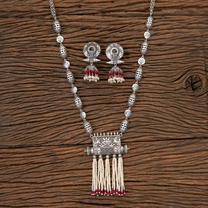 May include: A silver necklace with a rectangular pendant featuring intricate designs and a tassel of white beads with red accents. The necklace is paired with matching earrings with a circular design and red beads.