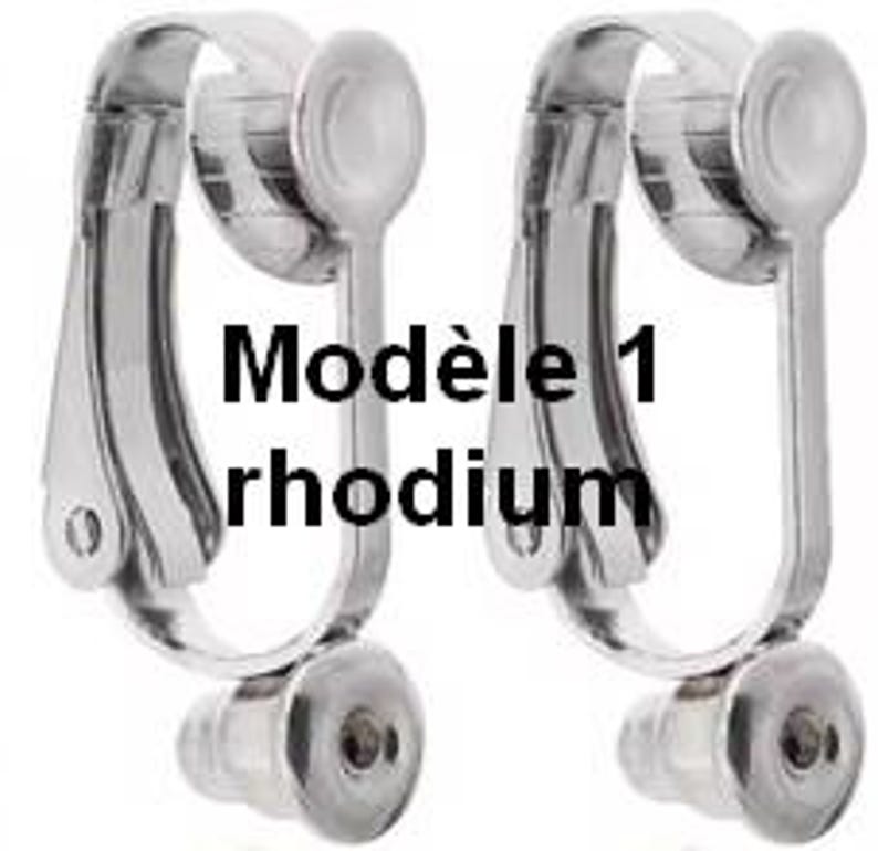 May include: A pair of silver-toned metal clip-on earring converters with a hinged mechanism and a circular base. The text "Mod&egrave;le 1 rhodium" is printed in black across the centre of the image, overlaying the two identical earring components.