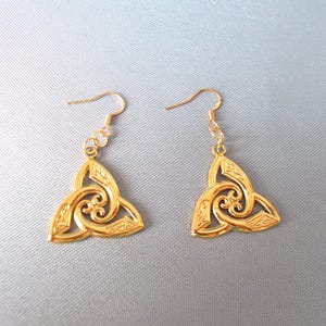 Puede incluir: Un par de pendientes de tono dorado con un diseño de nudo celta. Los pendientes están hechos de metal y tienen forma de gota.