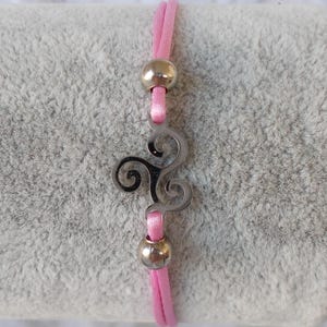 Könnte beinhalten: Rosa Kordelarmband mit einem silberfarbenen Triskele-Anhänger und zwei silbernen Perlen. Das Triskele ist ein keltisches Symbol mit drei ineinandergreifenden Spiralen. Das Armband liegt auf einer grauen, strukturierten Oberfläche.