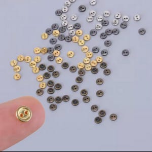 Pode incluir: Uma coleção de pequenos botões redondos de dois furos com acabamentos metálicos em dourado, prateado e cinza escuro. Os botões estão espalhados pelo quadro, com um botão dourado segurado em primeiro plano para mostrar sua forma circular.