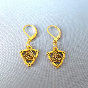 Puede incluir: Un par de pendientes dorados con cierre de palanca. Cada pendiente presenta un diseño de nudo celta dentro de un marco triangular, acentuado con pequeños detalles en forma de estrella. Los pendientes se muestran sobre un fondo azul claro.