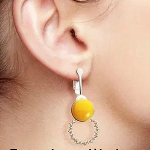 May include: A silver-toned clip-on earring featuring a smooth, round, yellow cabochon stone positioned above a circular, open-work frame adorned with small, clear, sparkling crystals. The earring is attached to a silver-coloured metal clip mechanism.