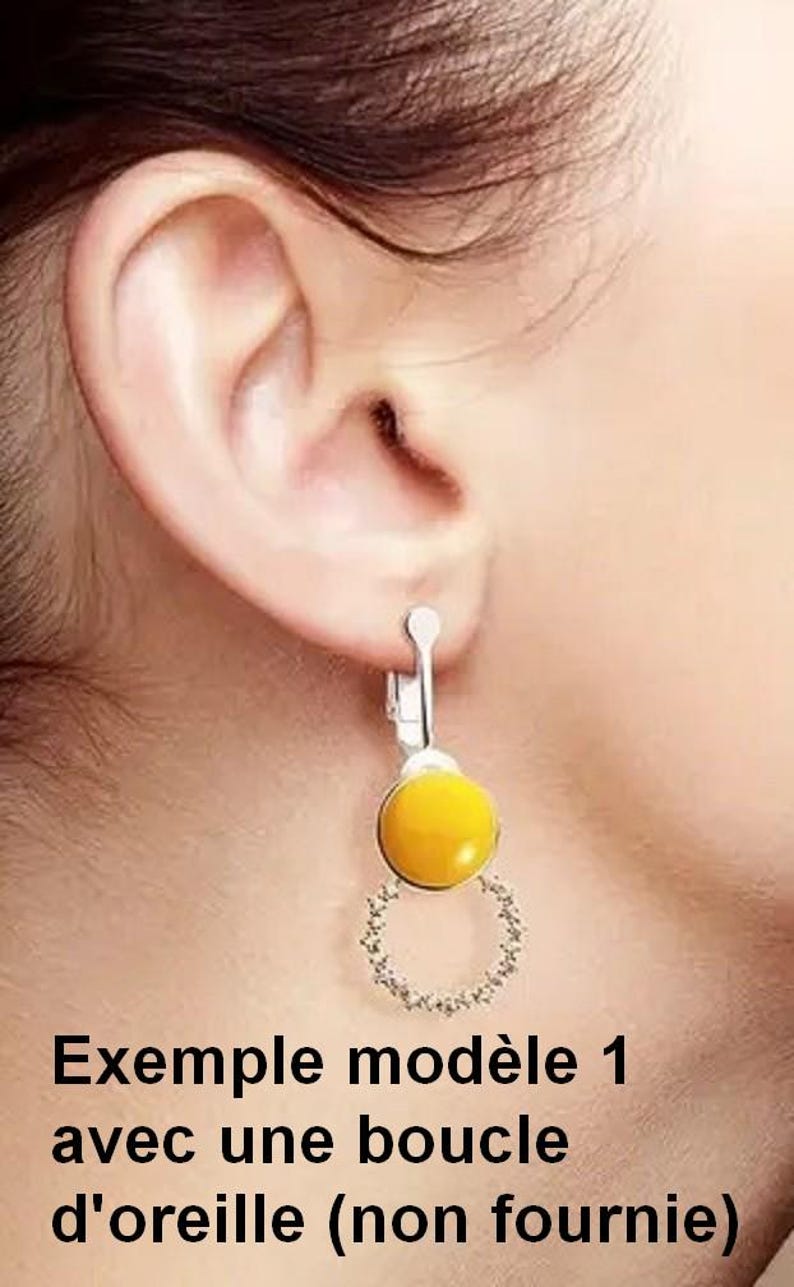May include: A silver-toned clip-on earring featuring a smooth, round, yellow cabochon stone positioned above a circular, open-work frame adorned with small, clear, sparkling crystals. The earring is attached to a silver-coloured metal clip mechanism.