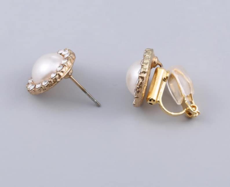 May include: A pair of earrings featuring a central white faux pearl surrounded by a halo of small clear crystals set in a textured gold-tone metal frame. One earring has a post back, while the other features a hinged clip-on mechanism.