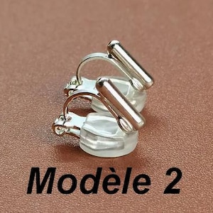May include: A pair of silver-toned clip-on earrings featuring a smooth, cylindrical bar design attached to a curved metal wire and a clear, rounded silicone cushion pad. The text "Mod&egrave;le 2" is printed in black below the earrings.