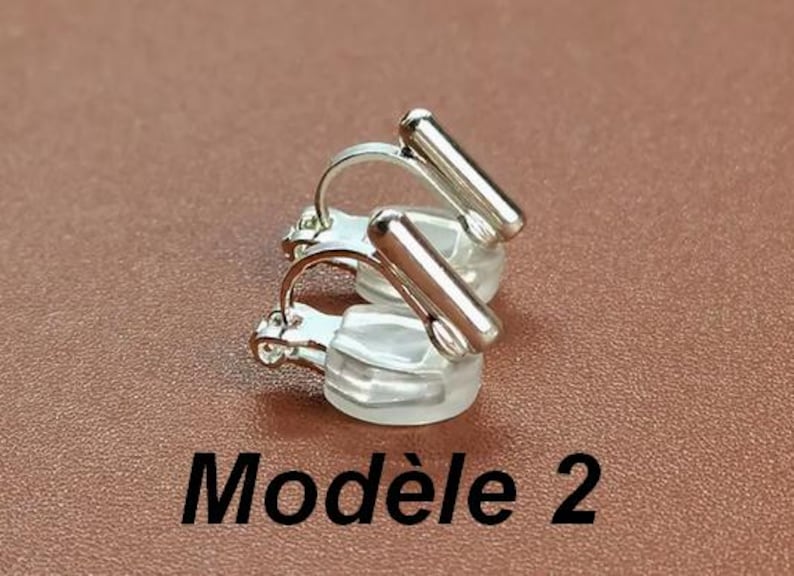 May include: A pair of silver-toned clip-on earrings featuring a smooth, cylindrical bar design attached to a curved metal wire and a clear, rounded silicone cushion pad. The text "Mod&egrave;le 2" is printed in black below the earrings.