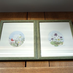 Könnte beinhalten: Zwei gerahmte Drucke von botanischen Illustrationen. Der linke Druck zeigt einen roten Schmetterling und weiße Blumen in einem kreisförmigen Design. Der rechte Druck zeigt lila Blumen in einem kreisförmigen Design. Beide Drucke haben einen grünen Rahmen.