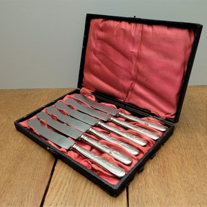 Könnte beinhalten: Ein Set mit sechs silbernen Buttermessern in einer schwarzen, mit Samt ausgekleideten Box. Die Messer haben verzierte Griffe und sind in einer Reihe angeordnet.