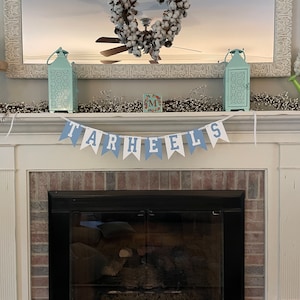 K&ouml;nnte beinhalten: Ein wei&szlig;er Kaminsims mit einem blau-wei&szlig;en Banner, auf dem "TARHEELS" steht. Das Banner h&auml;ngt &uuml;ber einem schwarzen Kamin mit einem Backsteinsockel. Auf dem Kaminsims stehen zwei t&uuml;rkisfarbene Laternen, ein Kranz mit Baumwollkugeln und eine kleine Holzkiste mit dem Buchstaben "M" darauf.