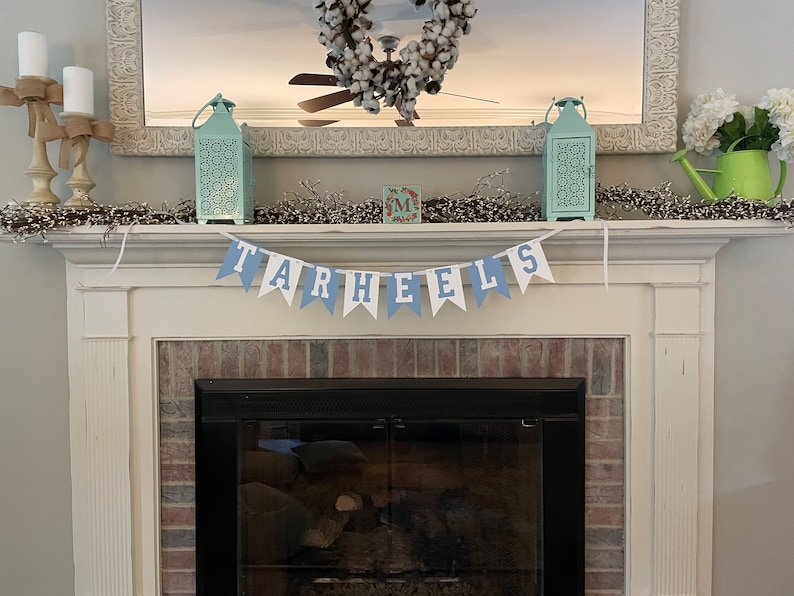 K&ouml;nnte beinhalten: Ein wei&szlig;er Kaminsims mit einem blau-wei&szlig;en Banner, auf dem "TARHEELS" steht. Das Banner h&auml;ngt &uuml;ber einem schwarzen Kamin mit einem Backsteinsockel. Auf dem Kaminsims stehen zwei t&uuml;rkisfarbene Laternen, ein Kranz mit Baumwollkugeln und eine kleine Holzkiste mit dem Buchstaben "M" darauf.