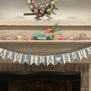 K&ouml;nnte beinhalten: Ein blaues und wei&szlig;es Banner mit dem Text "CAROLINA BOUND" in wei&szlig;en Buchstaben. Das Banner ist mit einer Abschluss-Kappe verziert und h&auml;ngt &uuml;ber einem Kamin.