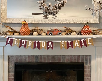 May include: A white fireplace mantel decorated with fall-themed decor, including a banner that reads "FLORIDA STATE" in white letters on a brown and burgundy background. The banner is strung with white ribbon and is flanked by two decorative gourds, one orange and one red and white.