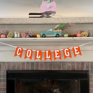 May include: Orange and white banner with the word "COLLEGE" on it. The banner is hanging on a white mantel above a fireplace. There are decorative items on the mantel, including a small blue truck, a pink bunny, and some greenery.
