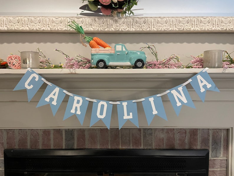 K&ouml;nnte beinhalten: Ein blaues und wei&szlig;es Banner mit dem Namen "CAROLINA" in wei&szlig;en Buchstaben auf blauem Hintergrund. Das Banner h&auml;ngt an einem wei&szlig;en Kaminsims &uuml;ber einem Kamin.
