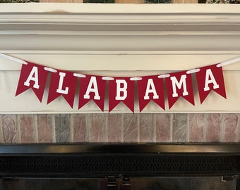 Puede incluir: Una pancarta roja y blanca con la palabra "ALABAMA" en letras blancas. La pancarta está colgada de una cuerda blanca y está enhebrada a través de una chimenea de ladrillo.