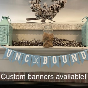 K&ouml;nnte beinhalten: Ein blaues und wei&szlig;es Banner mit dem Text "UNC x BOUND" und einer Grafik eines Hockeyschl&auml;gers. Das Banner h&auml;ngt an einem wei&szlig;en Kaminsims mit zwei t&uuml;rkisfarbenen Laternen und einem Einmachglas mit Sackleinen darauf. Der Text "Individuelle Banner verf&uuml;gbar!" befindet sich unter dem Banner.