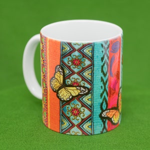 May include: A white ceramic mug with a colorful design featuring butterflies and geometric patterns in shades of orange, blue, green, and red.