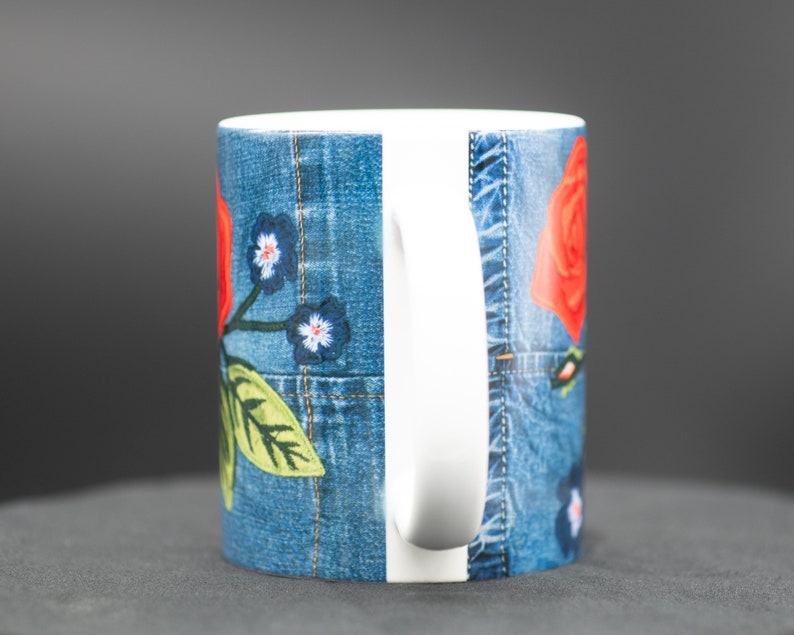 May include: A white ceramic mug with a denim-inspired design featuring red roses and blue flowers. The mug has a white handle.