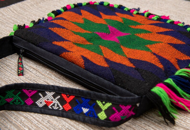 May include: A colorful woven bag with a geometric pattern in shades of orange, green, blue, and black. The bag has a zipper closure and a long strap with embroidered details.