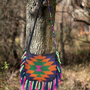 May include: A colorful woven bag with fringe hanging from the bottom. The bag is a dark blue with orange, green, and pink geometric patterns. The bag is hanging from a tree branch.