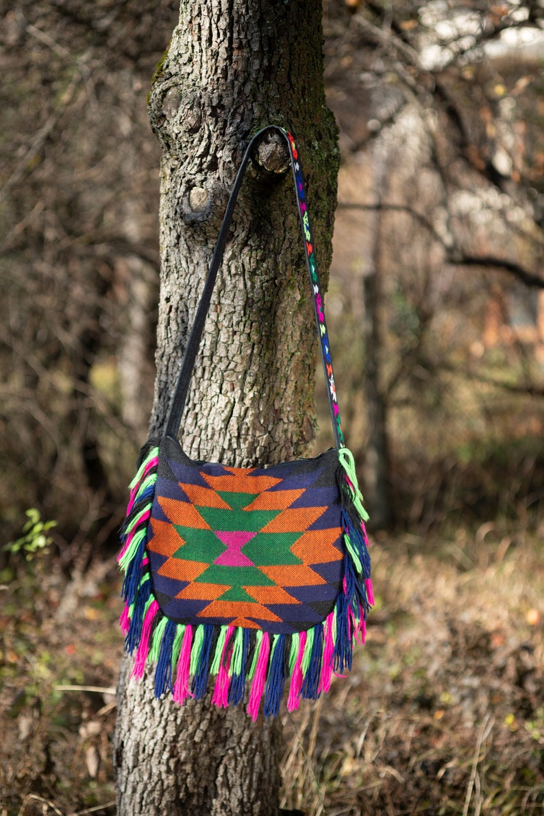 May include: A colorful woven bag with fringe hanging from the bottom. The bag is a dark blue with orange, green, and pink geometric patterns. The bag is hanging from a tree branch.