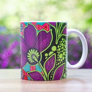 Pode incluir: Uma caneca de cer&acirc;mica branca com um design floral colorido. O design apresenta flores roxas, verdes e pretas sobre um fundo preto.