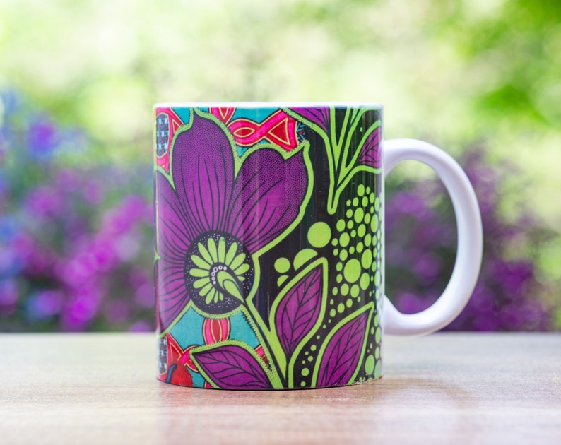 Pode incluir: Uma caneca de cer&acirc;mica branca com um design floral colorido. O design apresenta flores roxas, verdes e pretas sobre um fundo preto.