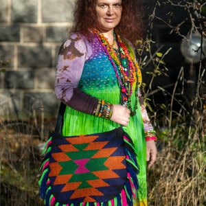May include: A woman wearing a colorful tie-dye dress with a beaded necklace and bracelets. She is carrying a large, colorful, fringed bag with a geometric pattern.