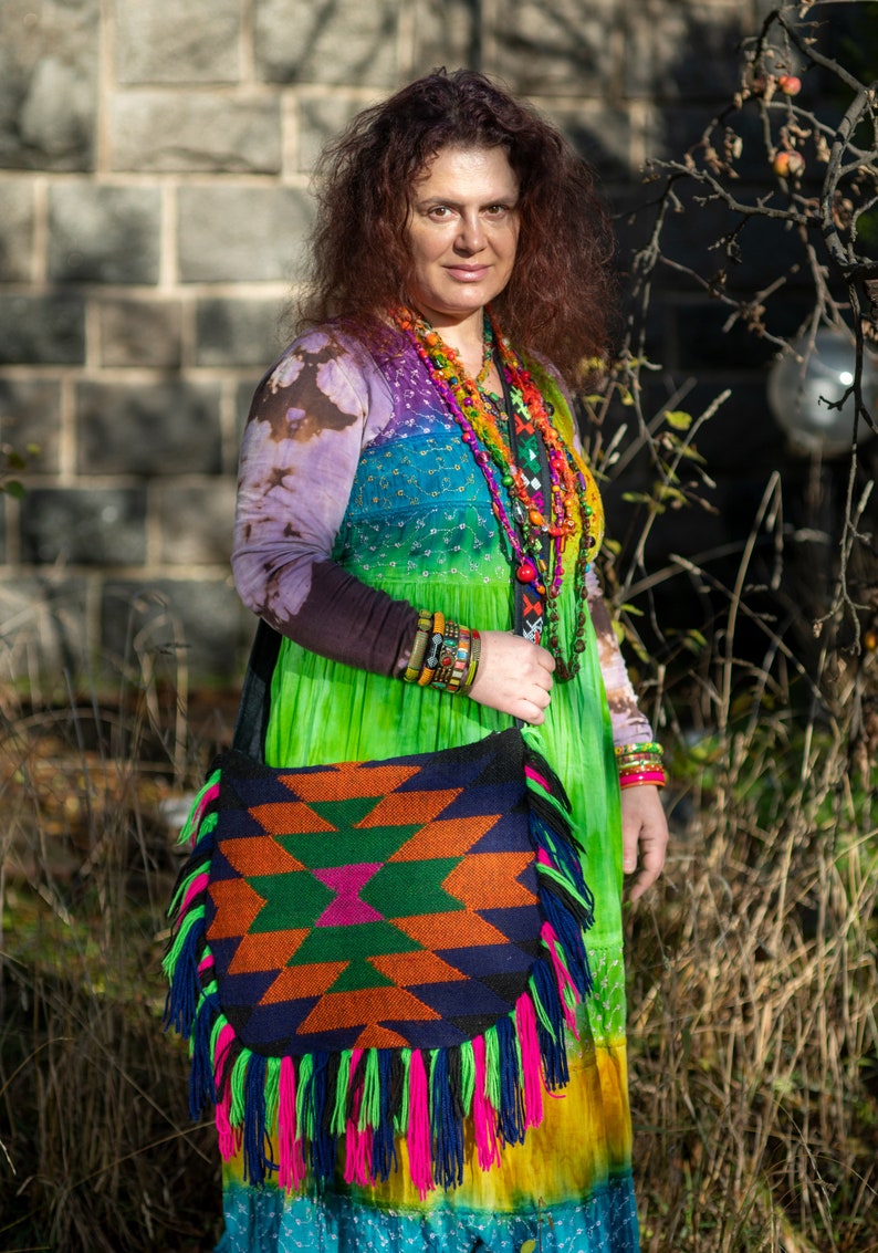 May include: A woman wearing a colorful tie-dye dress with a beaded necklace and bracelets. She is carrying a large, colorful, fringed bag with a geometric pattern.