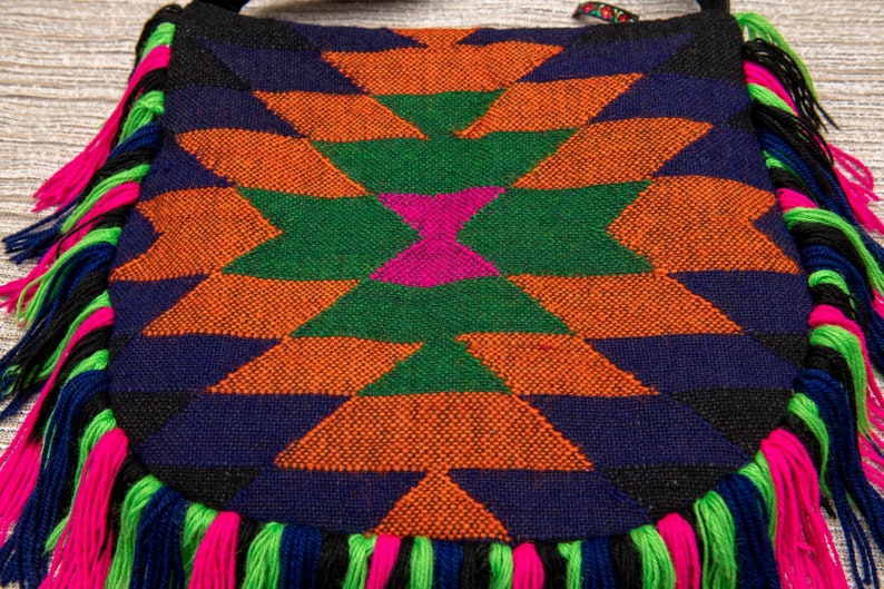 May include: A colorful woven bag with a geometric pattern in shades of orange, green, pink, blue, and black. The bag has a fringe border in pink, green, and blue.
