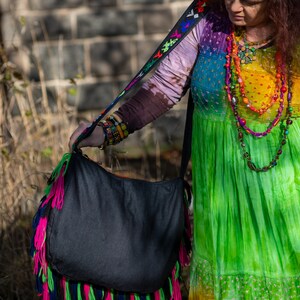 May include: A woman wearing a colorful tie-dye dress and a beaded necklace is holding a black fringed tote bag with a colorful strap.