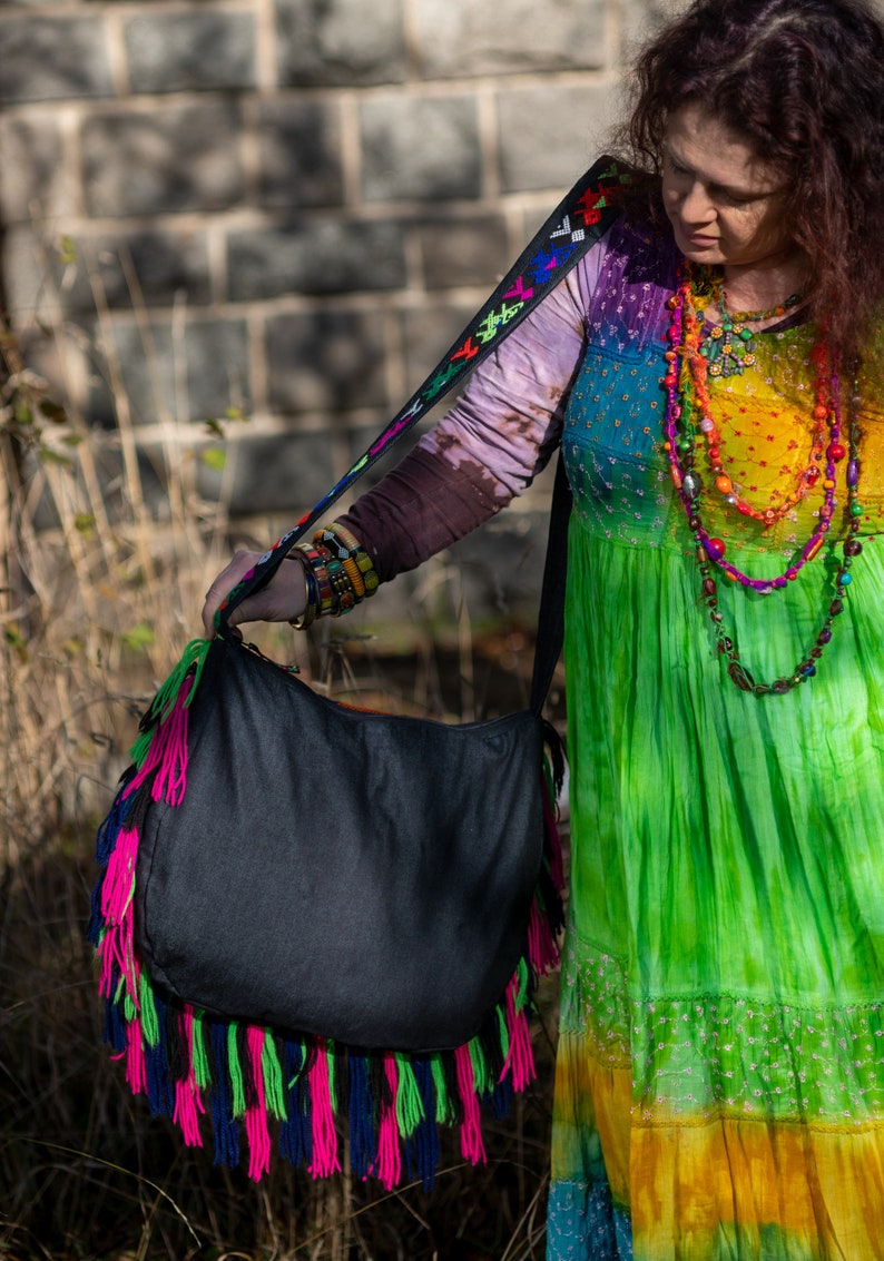 May include: A woman wearing a colorful tie-dye dress and a beaded necklace is holding a black fringed tote bag with a colorful strap.