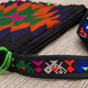 May include: A black crossbody bag with a colorful geometric pattern and fringe. The bag has a zipper closure and a black adjustable strap with a colorful embroidered trim.