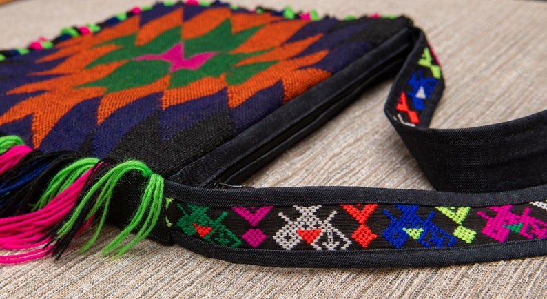 May include: A black crossbody bag with a colorful geometric pattern and fringe. The bag has a zipper closure and a black adjustable strap with a colorful embroidered trim.
