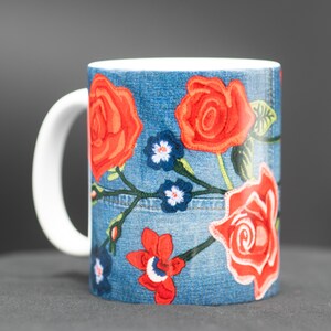 May include: A white ceramic mug with a blue denim pattern and red embroidered roses. The mug has a white handle.