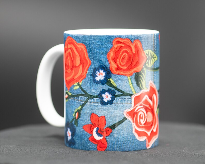 May include: A white ceramic mug with a blue denim pattern and red embroidered roses. The mug has a white handle.