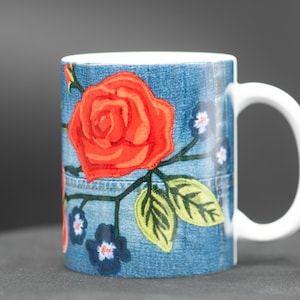 May include: A white ceramic mug with a blue denim pattern and red embroidered roses. The mug has a white handle.
