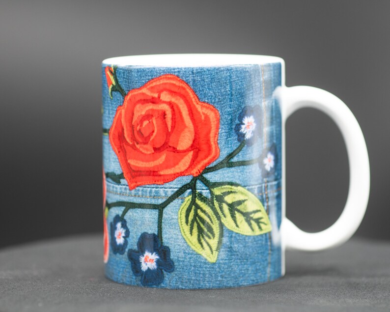 May include: A white ceramic mug with a blue denim pattern and red embroidered roses. The mug has a white handle.