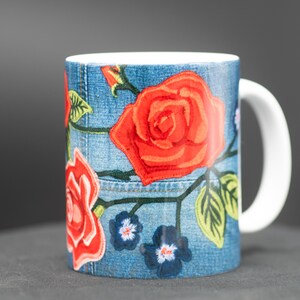 May include: A white ceramic mug with a blue denim pattern and red roses embroidered on it. The mug has a white handle.