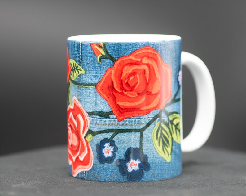 May include: A white ceramic mug with a blue denim pattern and red roses embroidered on it. The mug has a white handle.
